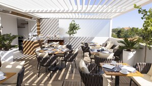An outdoor dining area with wooden tables and chairs, a pergola providing shade, and potted plants.