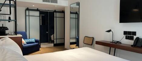 A hotel room with a bed, a desk with a computer, a chair, a sofa, a TV, and a bathroom with a shower and toilet.