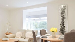 A modern living room with a sofa, dining table, and chairs. There is a window providing natural light and a view of greenery outside. - Princess Court Apartment (Llanelli)