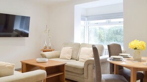 A modern living room with a sofa, a coffee table, a dining table with chairs, and a large window providing a view of greenery outside. - Princess Court Apartment (Llanelli)