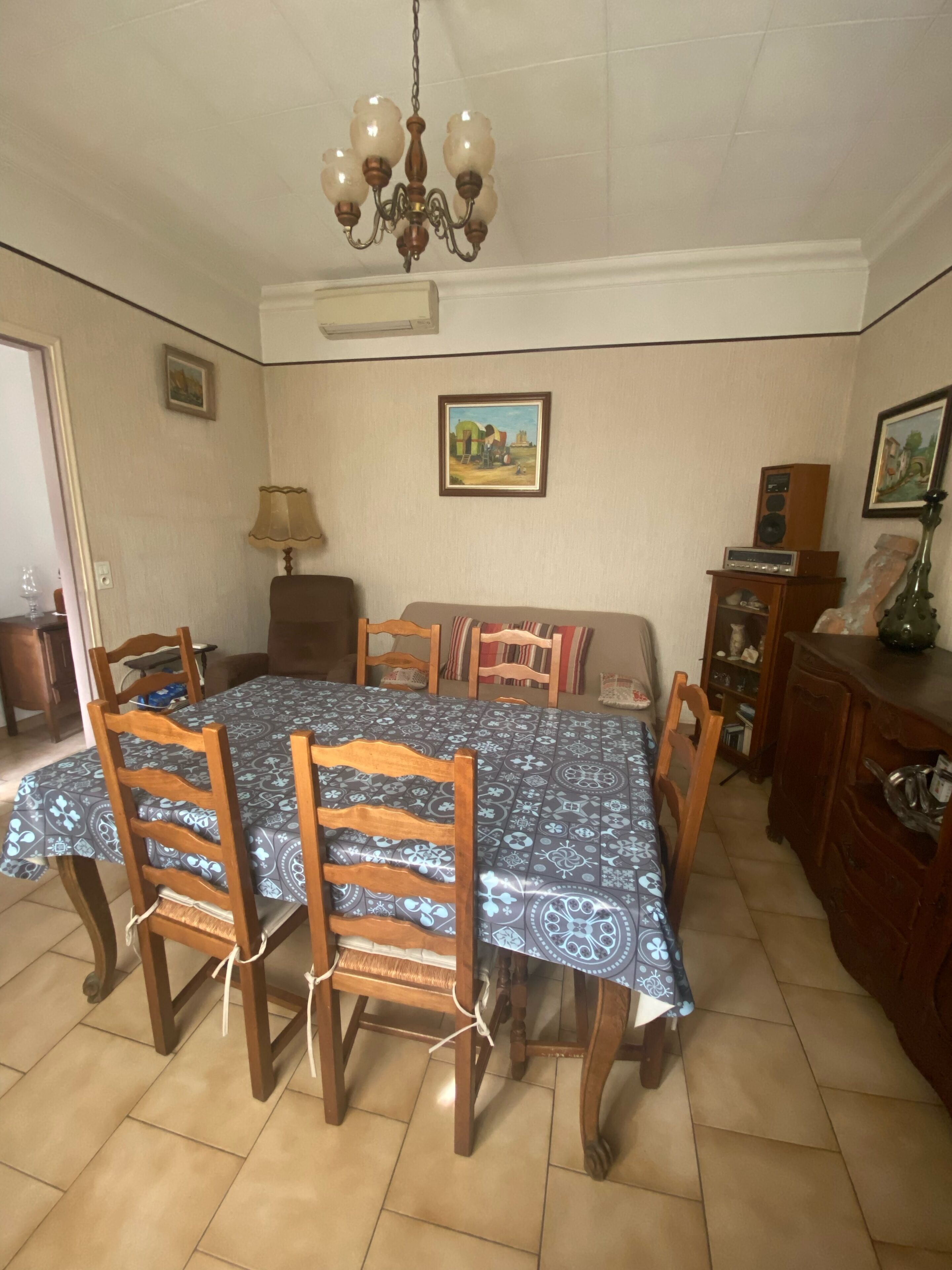 Une salle à manger avec une grande table recouverte d’une nappe à motifs, entourée de chaises en bois. Un lustre est suspendu au plafond et des tableaux encadrés ornent les murs.