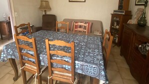 A dining room with a large table covered by a patterned tablecloth, surrounded by wooden chairs. A chandelier hangs from the ceiling, and there are framed pictures on the walls.