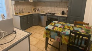 A compact kitchen with grey cabinets, a tiled backsplash, a dining table with a colorful tablecloth, and a hanging light fixture.