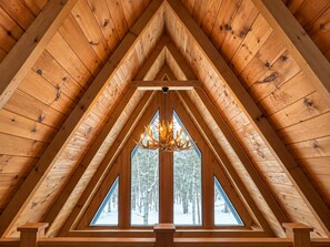 Un dormitorio rústico en el ático con techo inclinado, una araña y vistas a un bosque nevado a través de amplios ventanales.