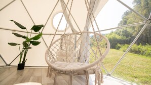 Une chaise suspendue en macramé blanc se trouve dans une pièce dotée d’une grande fenêtre, d’une plante en pot et d’un plancher en bois.