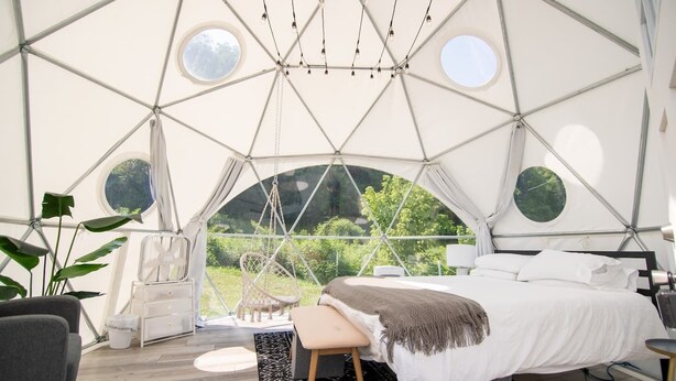 Une pièce aménagée comme une tente, avec un lit, une chaise, un ventilateur et une vue sur la verdure environnante.