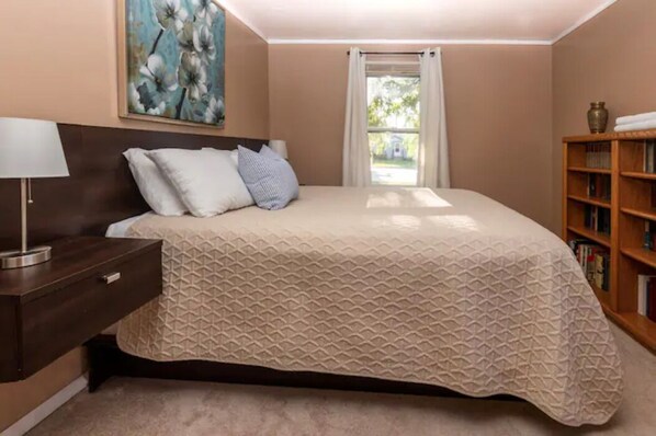 A bedroom with a bed, a nightstand, a lamp, a bookshelf, and a window with curtains.
