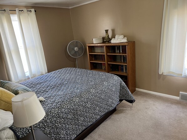 Une chambre à coucher avec un lit, une lampe de chevet, une bibliothèque et un ventilateur sur pied.