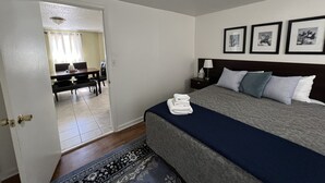 A hotel room with a bed, bedside table, lamp, and a view into the dining area.