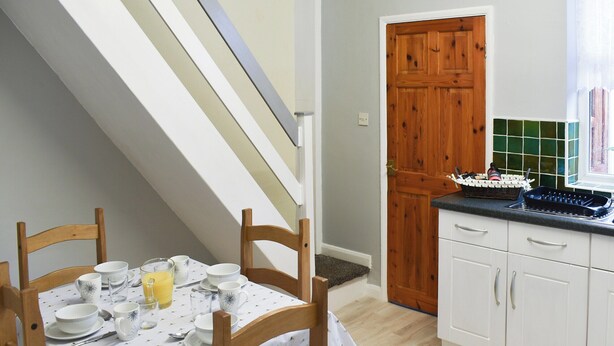 Un comedor con una mesa lista para el desayuno, una silla de madera, y una cocina con armarios blancos y una puerta de madera.