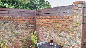 Un muro de ladrillo con un portón de madera, una mesa con dos copas de vino y dos macetas con plantas.