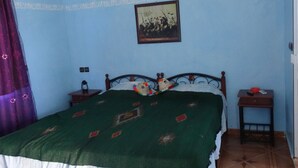 Un dormitorio con cama, mesita de noche, una pequeña mesa, un cuadro enmarcado en la pared y una cortina.