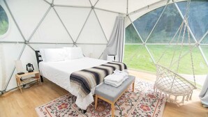 Une chambre aménagée comme une tente, avec un lit, un banc, un fauteuil suspendu et une vue sur l’extérieur.