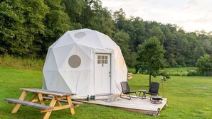 Une tente en forme de dôme géodésique dotée d’une porte et de fenêtres, ainsi qu’une table de pique-nique, deux chaises et une petite terrasse.