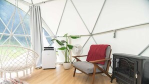 Une pièce chaleureuse avec un hamac, un fauteuil recouvert d’une couverture rouge, un ventilateur, un radiateur et une cheminée.