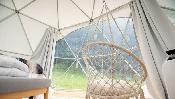 Une pièce chaleureuse avec un fauteuil suspendu, un lit et une grande fenêtre donnant sur un paysage verdoyant.