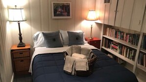 A neatly made bed with a blue comforter and pillows, a bedside lamp, a nightstand with a lamp, a bookshelf with books, and a framed picture on the wall. - Cozy English Basement Apt quiet but close to downtown (Winchester)