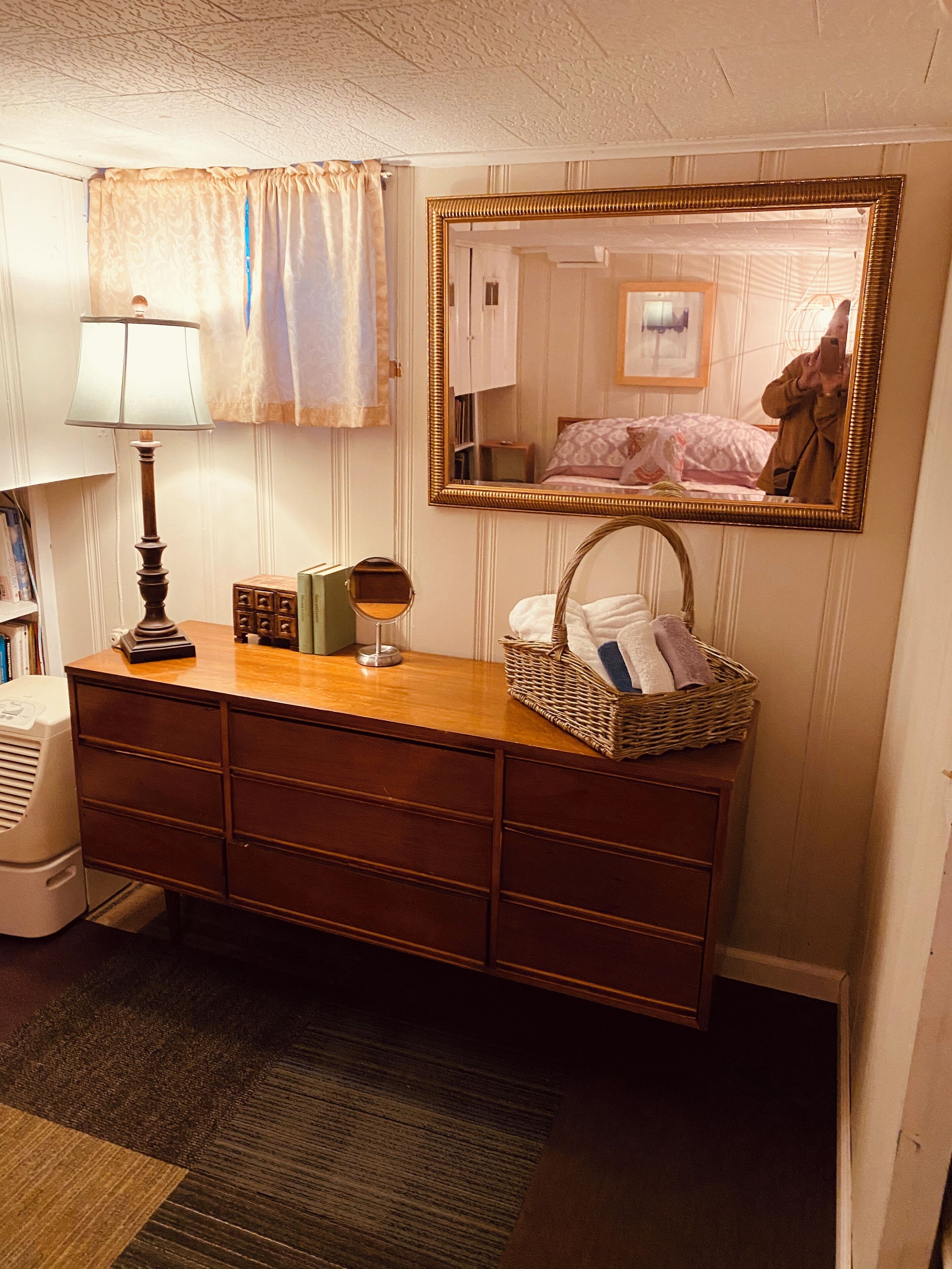 A bedroom with a wooden dresser, a mirror, a lamp, and a wicker basket with towels.