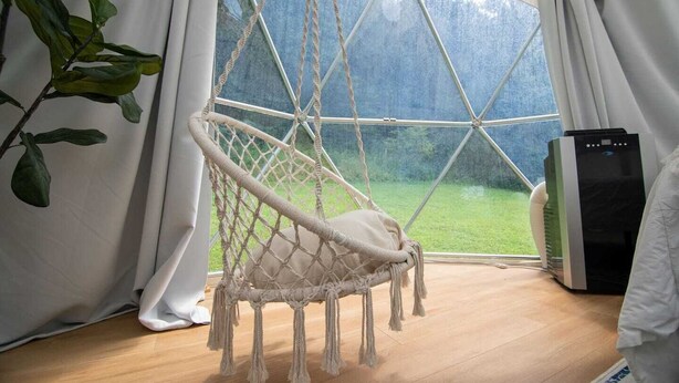 A room with a macrame hanging chair, a large window, and a black heater.