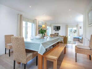 A dining area with a long wooden table, wicker chairs, and a bench. There is a television mounted on the wall and a window with curtains.