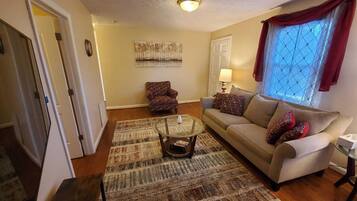 A living room with a beige sofa, patterned rug, and a coffee table. There is a window with red curtains and a painting on the wall.