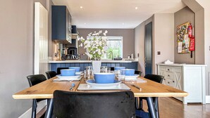 Un espace repas moderne avec une table en bois dressée pour six personnes, entourée de chaises bleu foncé. On aperçoit à l’arrière-plan une cuisine avec des meubles sombres et un plan de travail blanc.