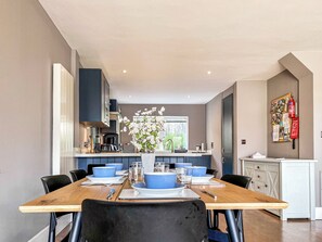 A modern dining area with a wooden table set for six, flanked by dark blue chairs. A kitchen with dark cabinets and a white countertop is visible in the background. - 4 bedroom accommodation in South Cerney (South Cerney)