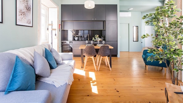 A modern living room with a sofa, dining table, and chairs. There is a kitchen in the background with dark cabinets and a black countertop.
