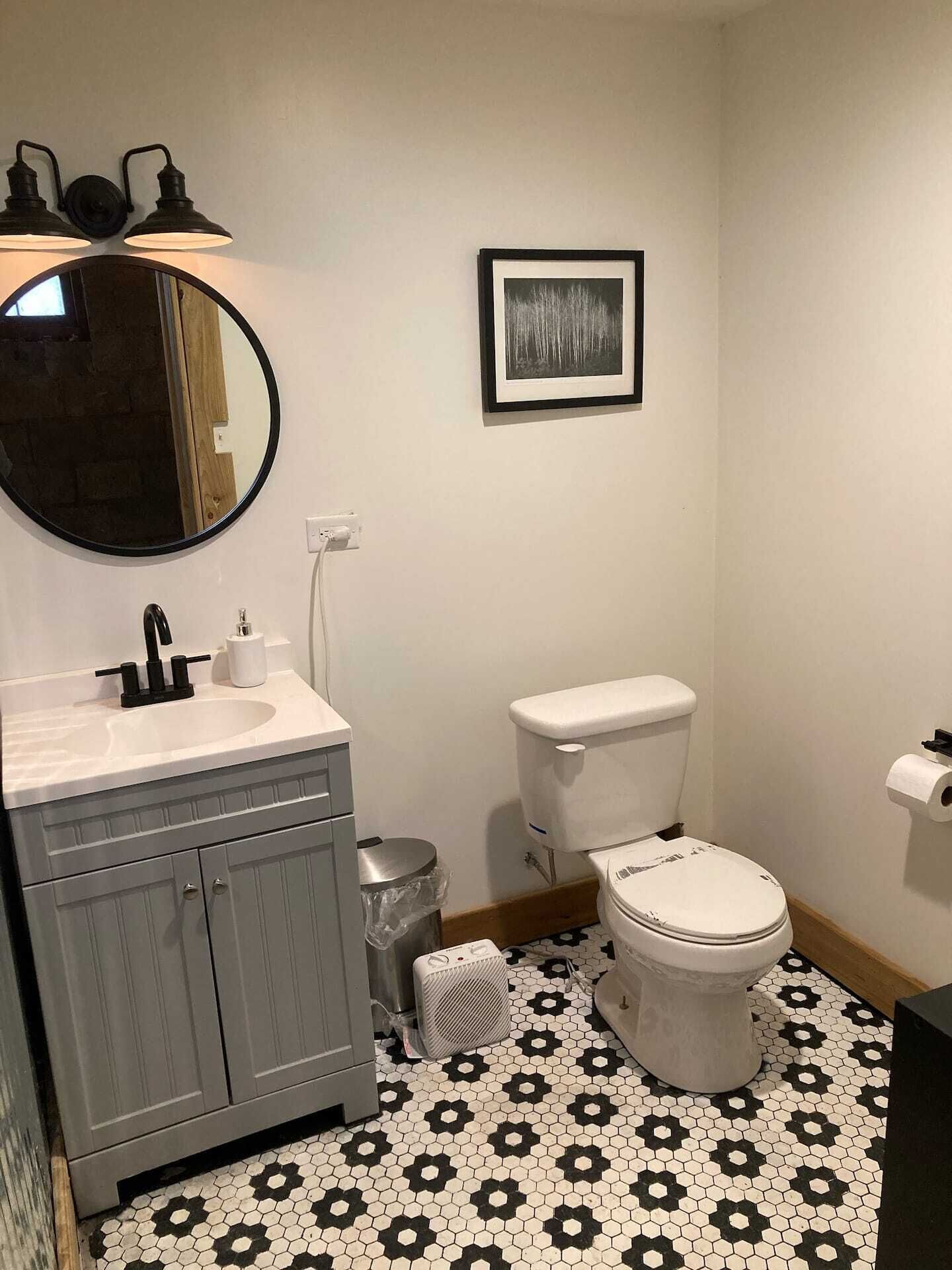 Ein Badezimmer mit einem weißen Waschbecken, einem schwarzen Wasserhahn, einem runden Spiegel, einer Toilette und einem gerahmten Bild an der Wand.