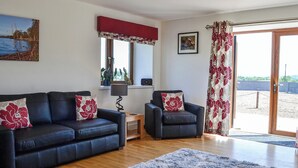 A living room with a dark blue sofa, a smaller armchair, a fluffy rug, a wooden coffee table, a lamp, and a window with a view of a body of water and greenery. - The Byre @ Camp Douglas (Castle Douglas)