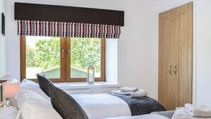 A bedroom with two beds, a window with a view of greenery, a wooden wardrobe, and a bedside table with a lamp. - The Byre @ Camp Douglas (Castle Douglas)
