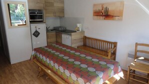 Cocina compacta con refrigerador, horno y cafetera. Hay una cama con una manta colorida y un banco de madera.