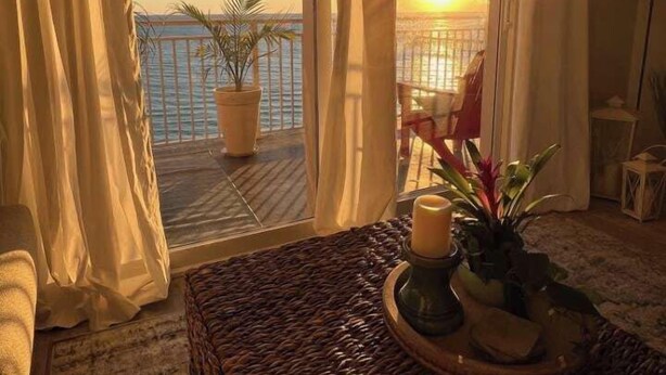 Un balcon avec vue sur le coucher de soleil sur la mer, une table en osier avec des bougies et une plante en pot.