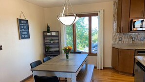 A dining area with a wooden table, chairs, a hanging light fixture, a view of the outdoors, and a kitchen with wooden cabinets and a microwave.