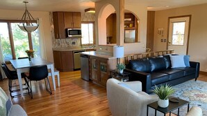 A modern living room with a black leather sofa, a dining table with chairs, a kitchen area with wooden cabinets, and a large window providing natural light.