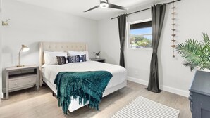 Un dormitorio con una cama, una mesita de noche, una lámpara, un ventilador de techo y una ventana con cortinas.