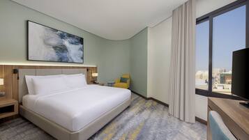 Habitación de hotel con una cama grande, un escritorio con silla, una mesita con lámpara, un cuadro en la pared y una ventana con vista a la ciudad.
