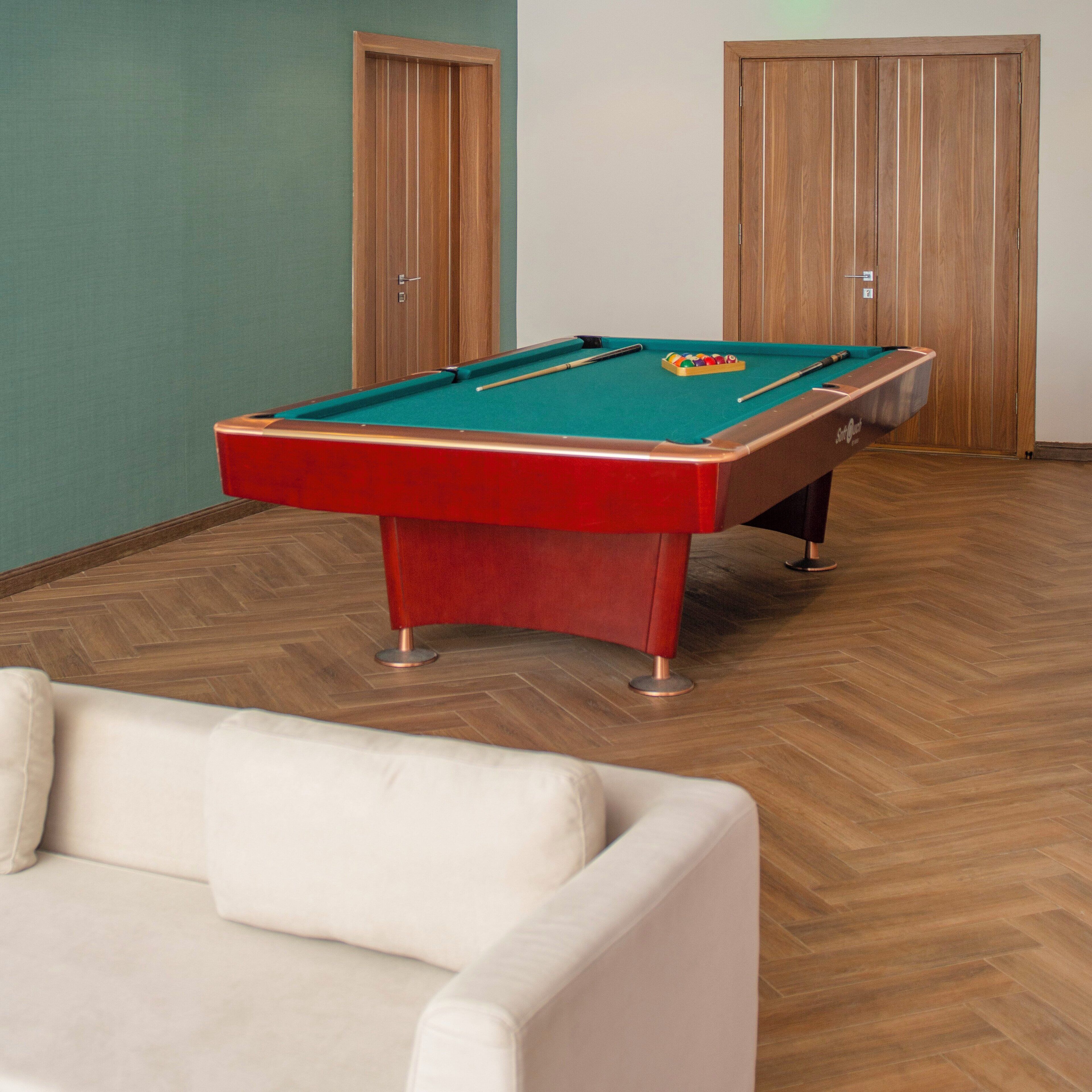 A pool table in a room with herringbone-patterned wooden flooring, a white sofa, and two wooden doors.