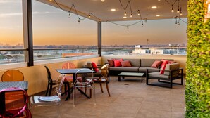 Un salon moderne situé sur un toit-terrasse, offrant une vue sur le paysage urbain, avec différents types d’espaces de détente et une table en verre.