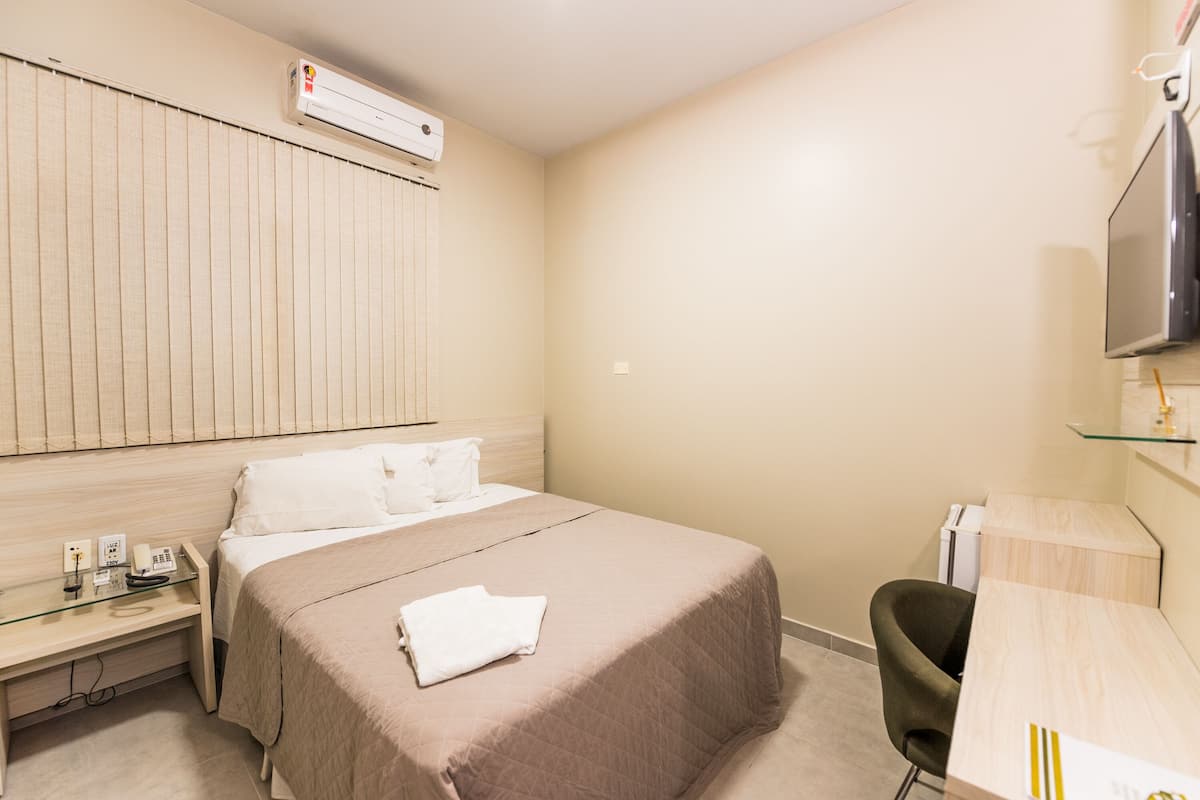 A hotel room with a bed, a desk, a chair, a television, and an air conditioning unit.