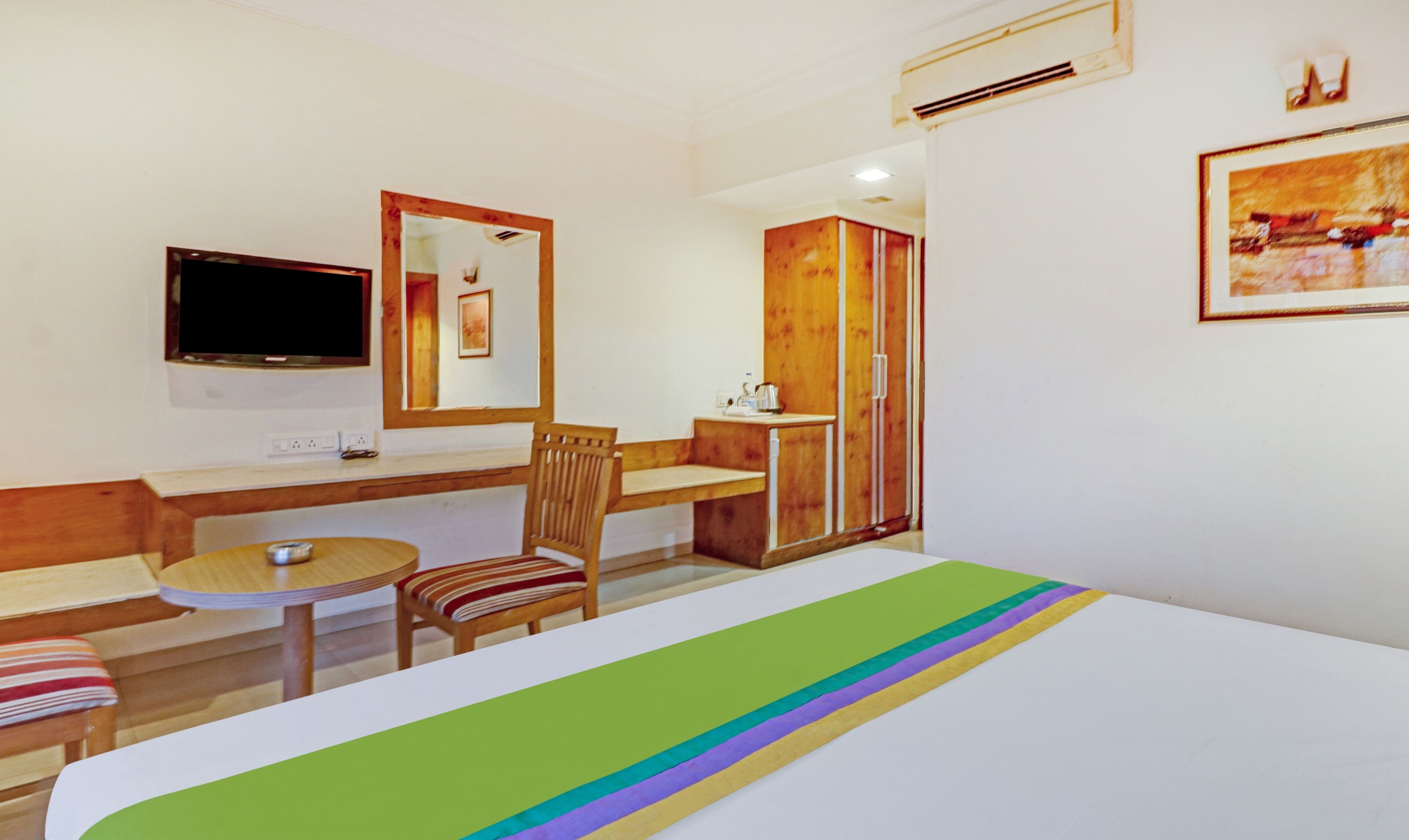 A hotel room with a bed, a desk, a chair, a small table, a television, a mirror, and a wardrobe.
