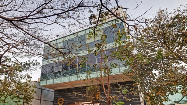 Um edifício moderno com fachada de vidro, uma placa com o nome Suraksha Inn e árvores ao redor.
