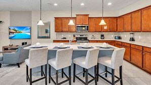A modern kitchen with wooden cabinets, a central island with bar stools, a microwave, oven, and a television displaying a tropical scene. - Dazzling 9BR Villa with Pool& Spa, Close to Disney (Four Corners)