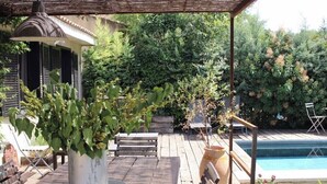 A wooden deck with a table and chairs, a potted plant, a pergola with a lamp, and a swimming pool.
