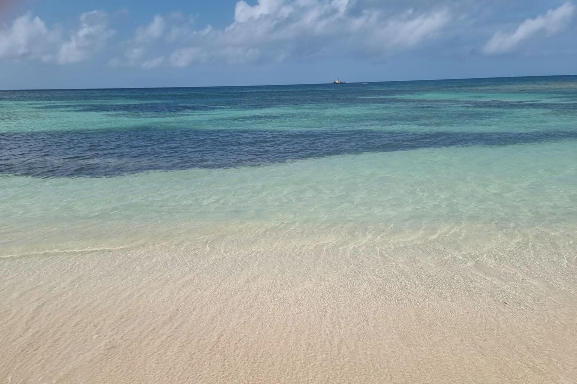 Uma praia com águas cristalinas e rasas, e um navio ao longe no horizonte.