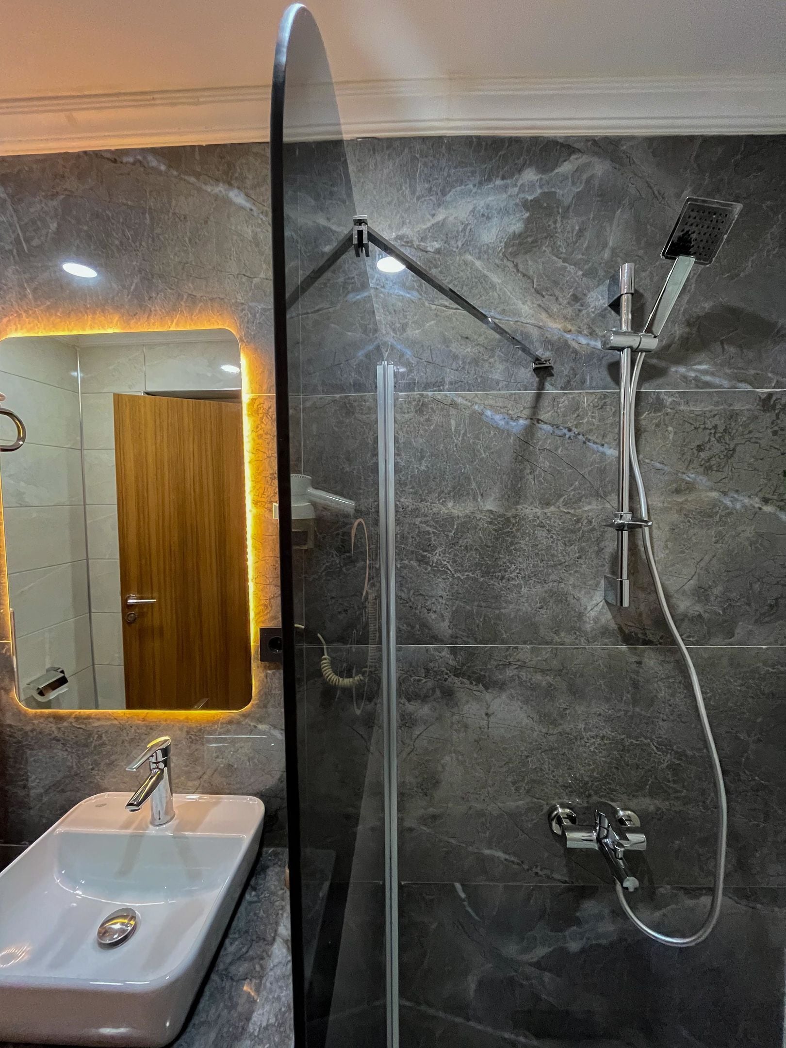 Een moderne badkamer met een douchecabine van glas, een wand betegeld met marmer, een houten deur en een wastafel met een chromen kraan.
