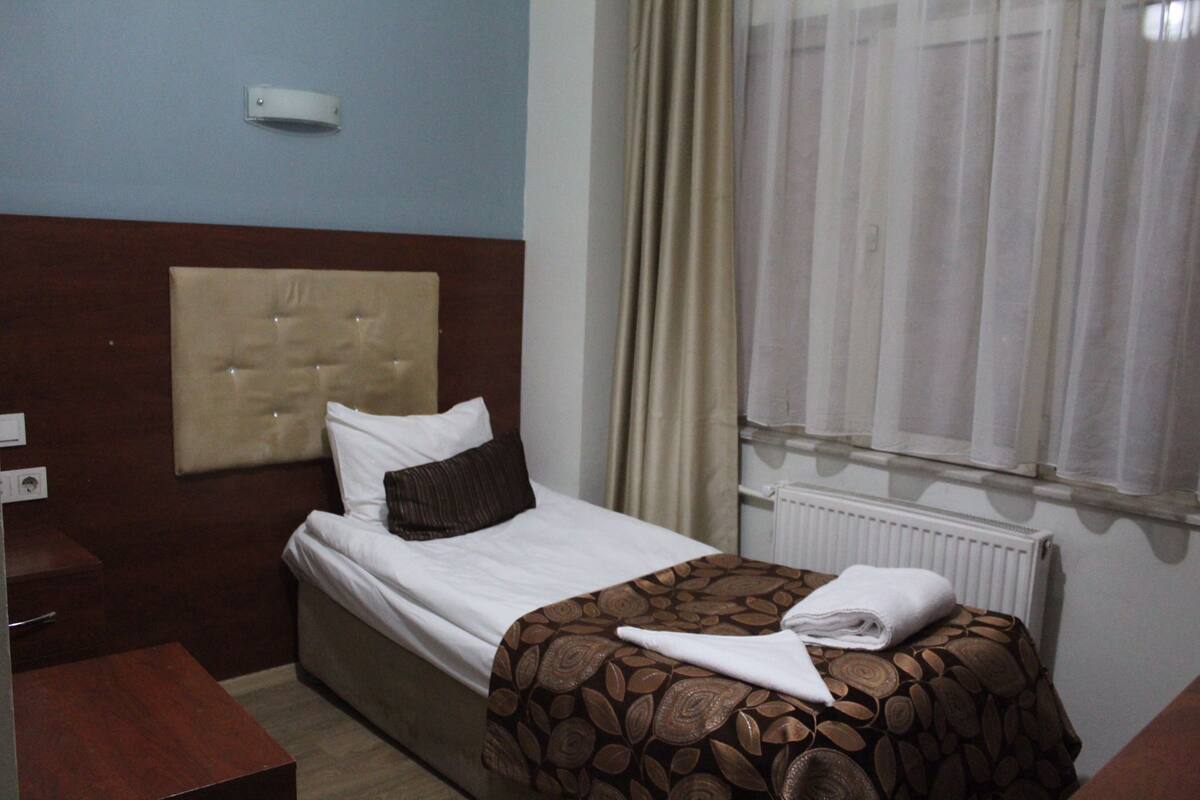 A hotel room with a bed, a bedside table, a window with curtains, and a wall-mounted light.