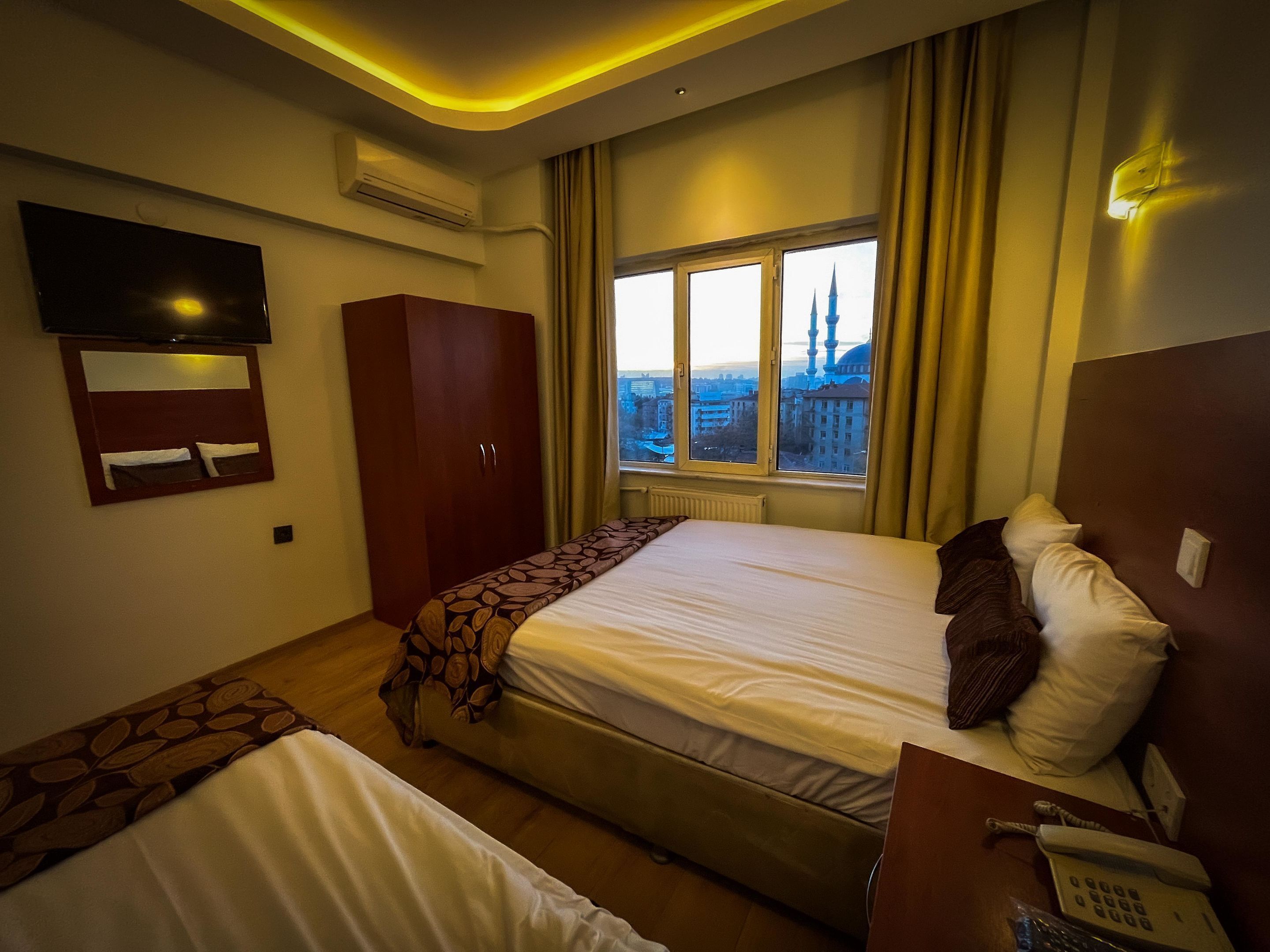 Quarto de hotel com duas camas, televisão, guarda-roupa, janela com vista para a cidade e mesa de cabeceira com telefone.