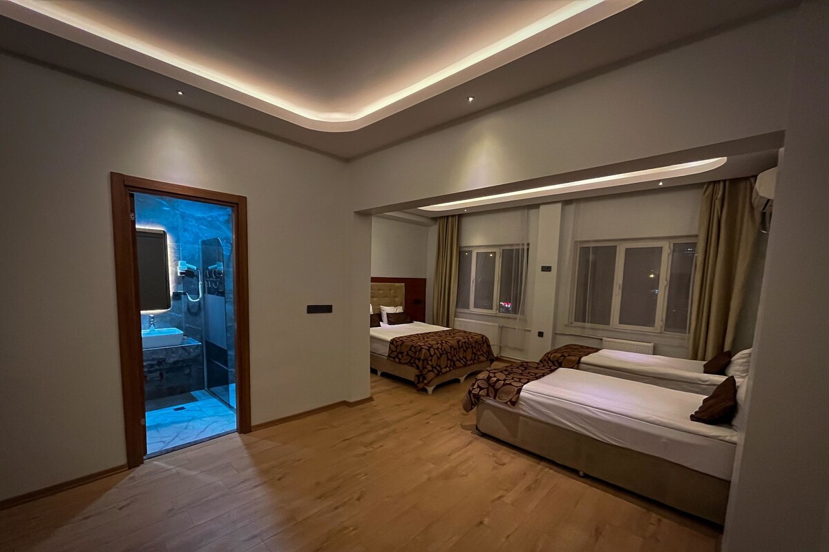 A hotel room with a wooden floor, a bed with patterned bedding, a bedside table, and a window with curtains.