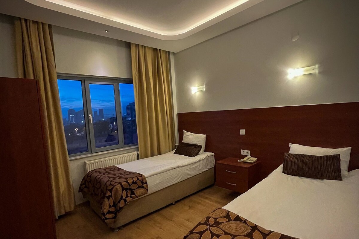 A hotel room with two beds, a window with curtains, a nightstand, and a wall-mounted light.
